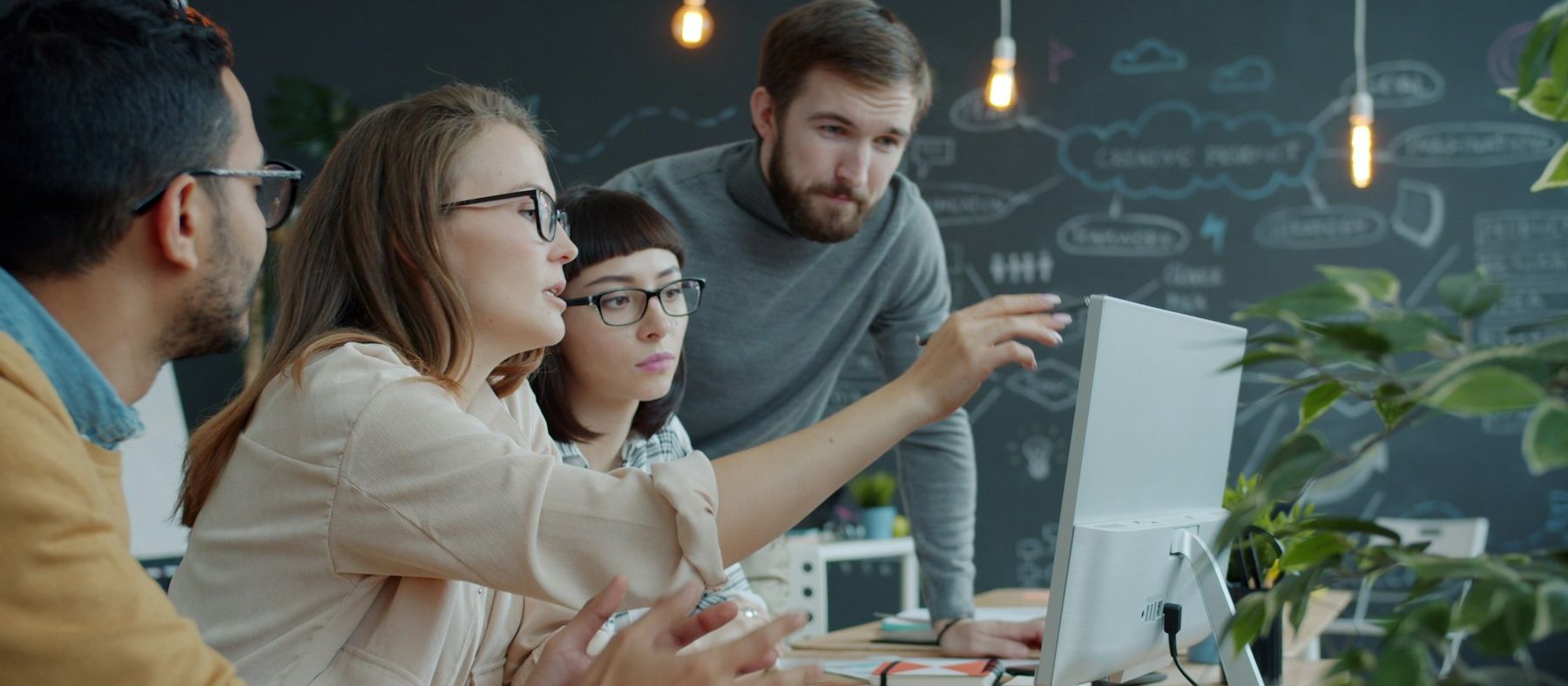 Four colleagues gathered around one screen in an open office, one pointing at it, a chalkboard of hand-drawn diagrams behind them.
