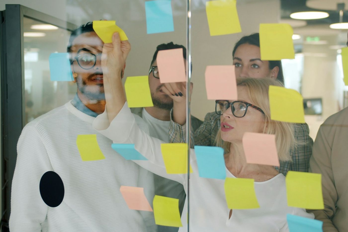 A team planning together with coloured sticky notes on a glass wall, one person reaching up to place a note.