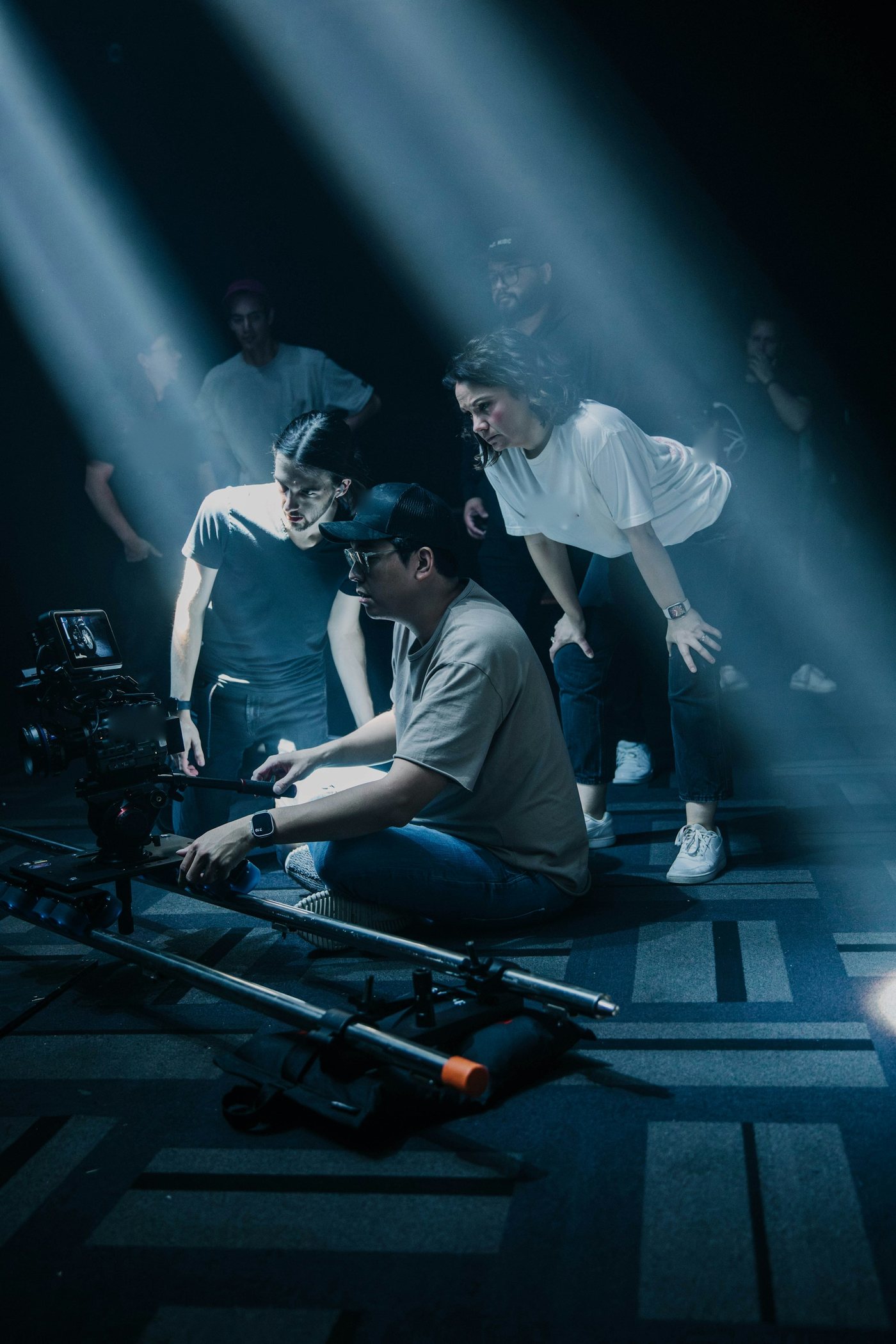 A camera operator at a slider in a beam of light, two crew members leaning in to watch the frame