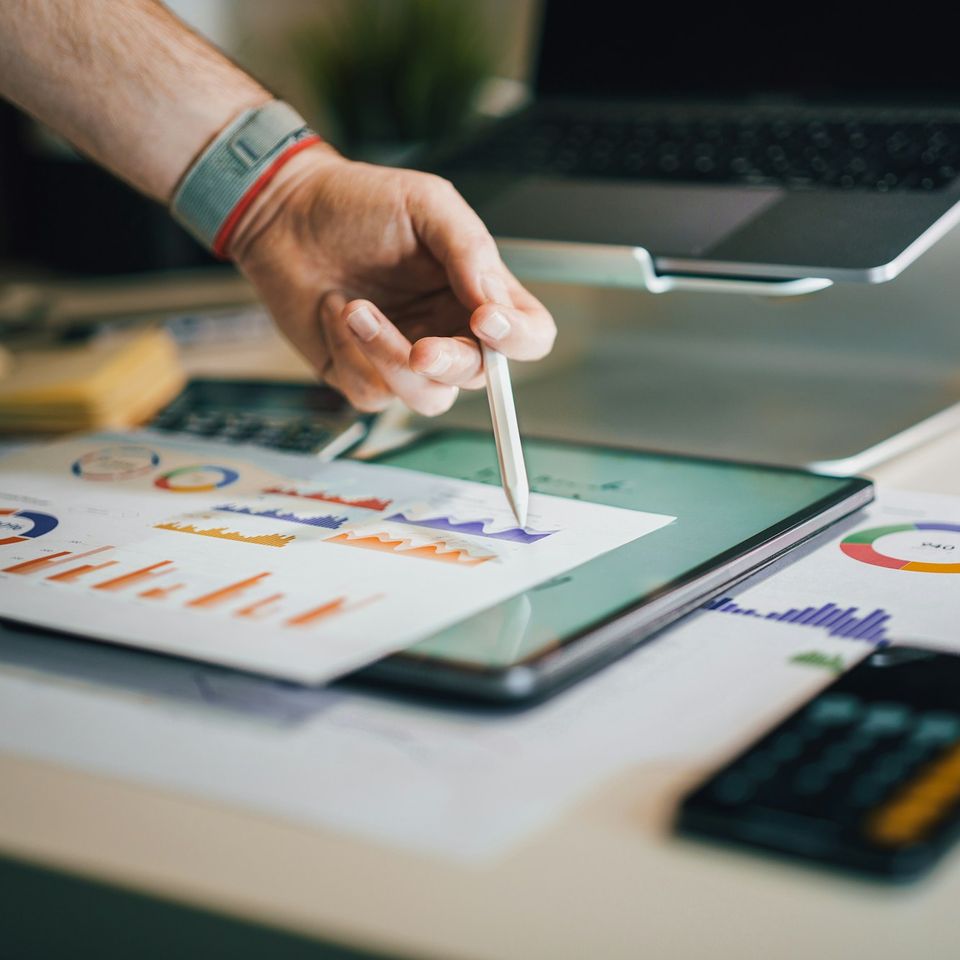 A hand with a stylus pointing at printed charts beside a tablet.
