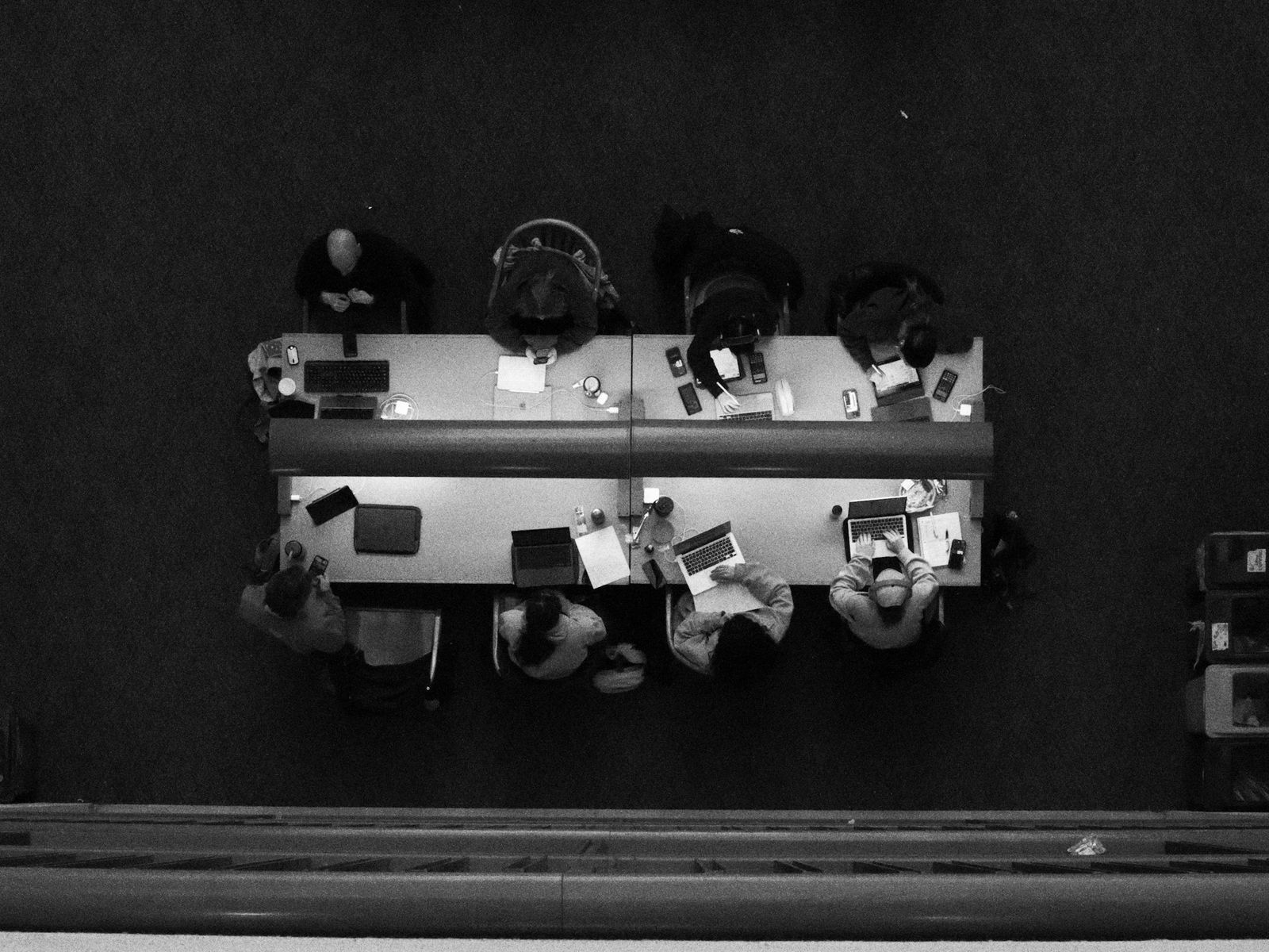 People working side by side at a long table, seen from above, in black and white.