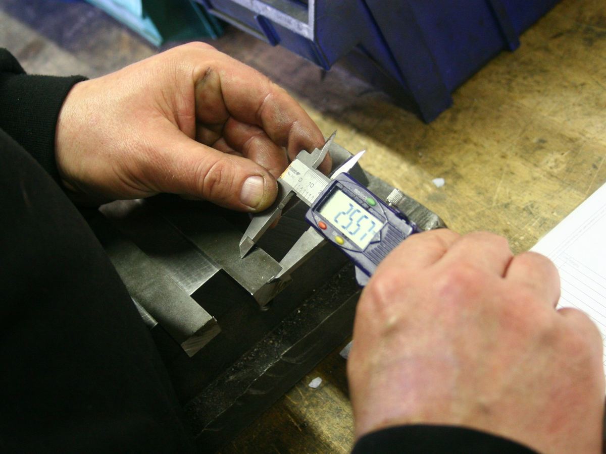 Hands measure a machined metal part with a digital caliper on a workshop bench, an inspection sheet beside it