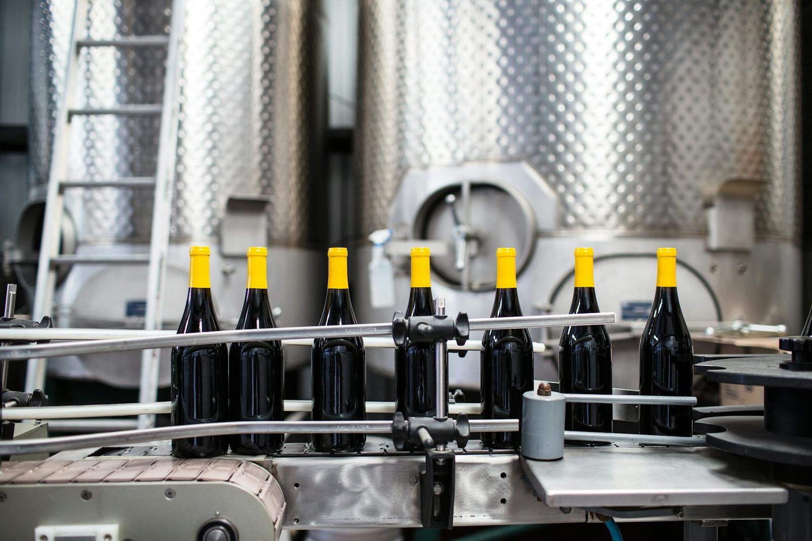 Seven dark wine bottles with yellow capsules pass along a conveyor in front of stainless steel tanks