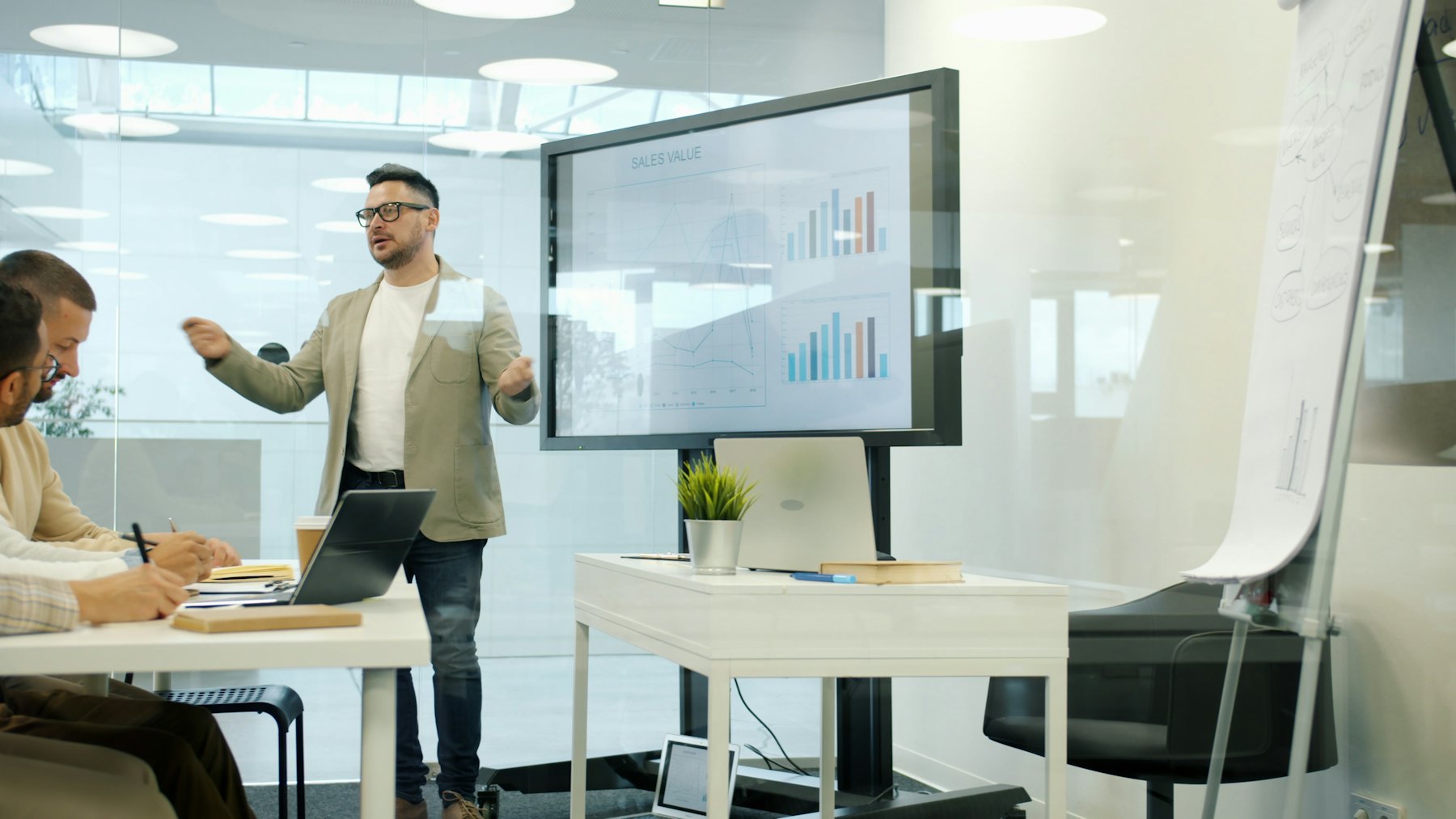 A presenter walks colleagues through a chart on a large screen in a glass-walled meeting room