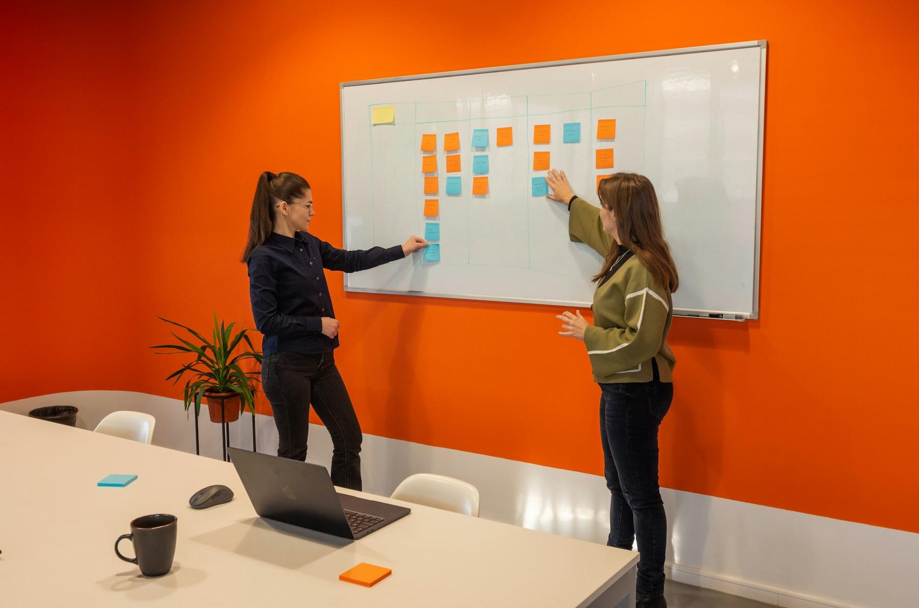 Two colleagues arrange sticky notes into columns on a whiteboard in a meeting room
