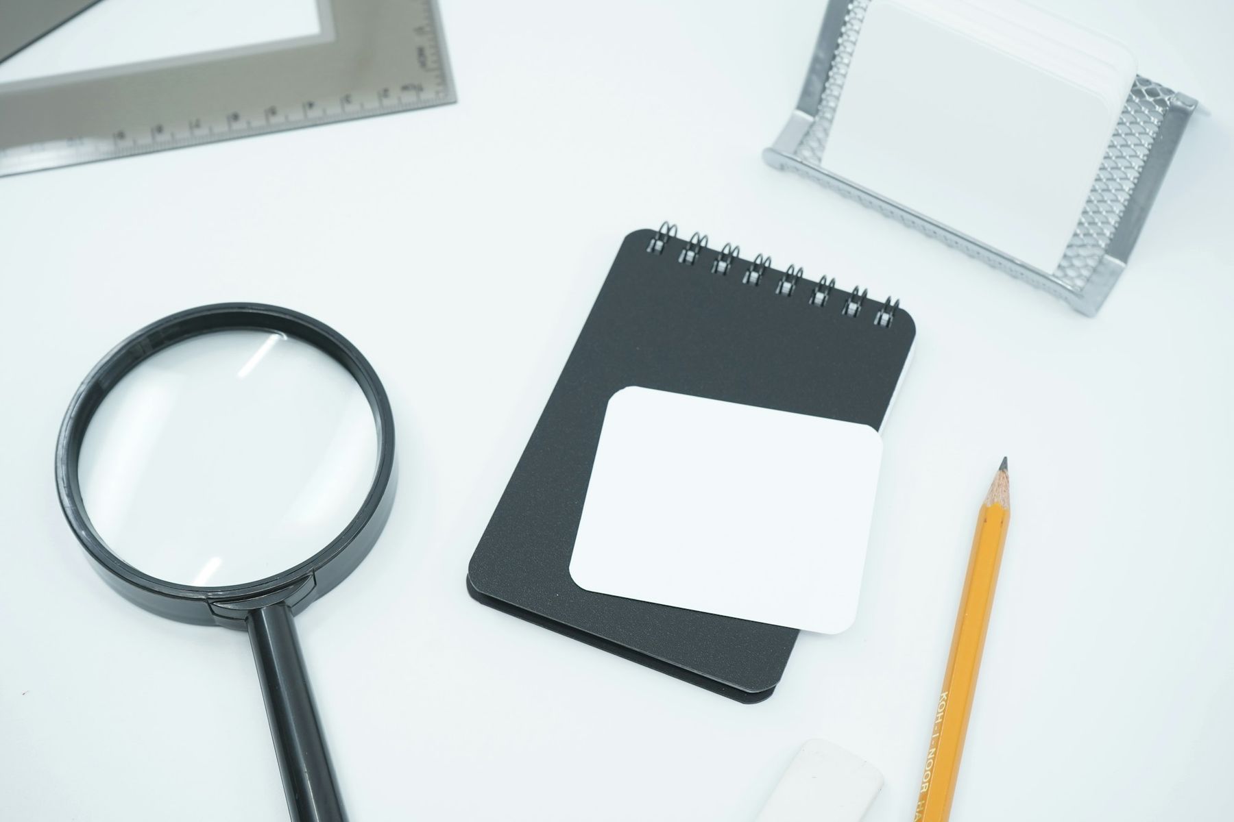 A black magnifying glass beside a small notebook and a pencil on a white desk