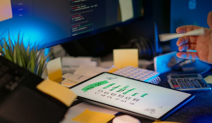 A desk with a monitor of source code, a handwritten list on a tablet, sticky notes and a calculator