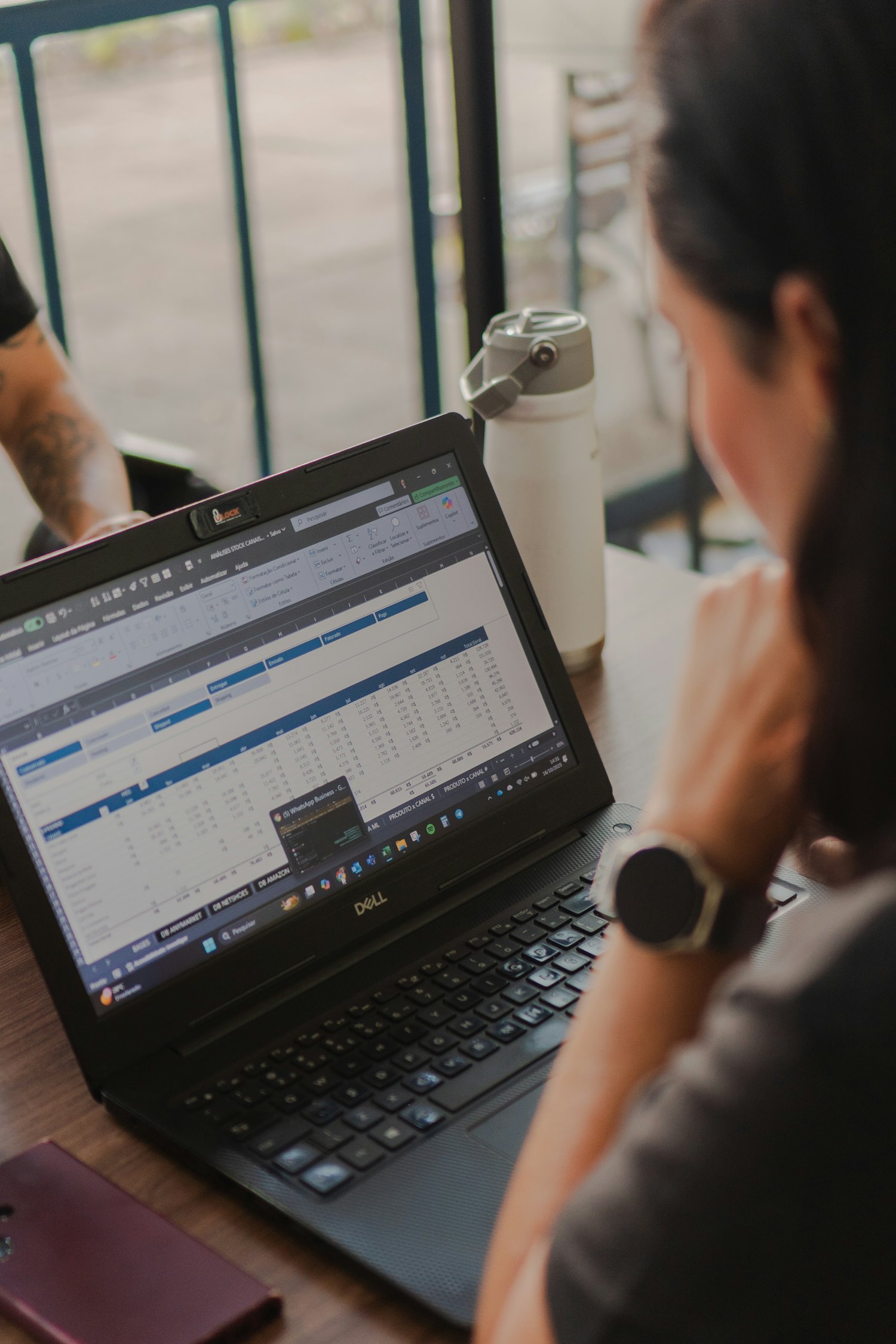 Over a colleague's shoulder: a laptop open on a dense spreadsheet of rows and columns at a shared table