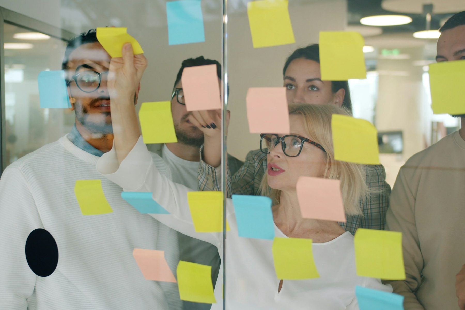 Five colleagues stand at a glass wall covered in sticky notes in a daylit office; one of them writes on a note while the others read the wall
