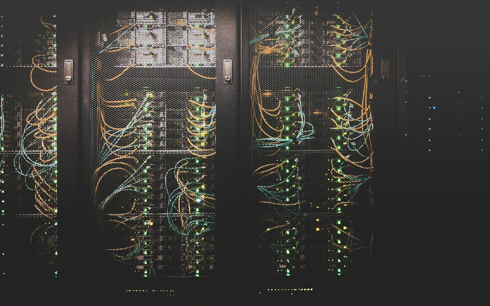 Rows of open server racks in a dark equipment room, orange and teal network cables looping past banks of green status lights