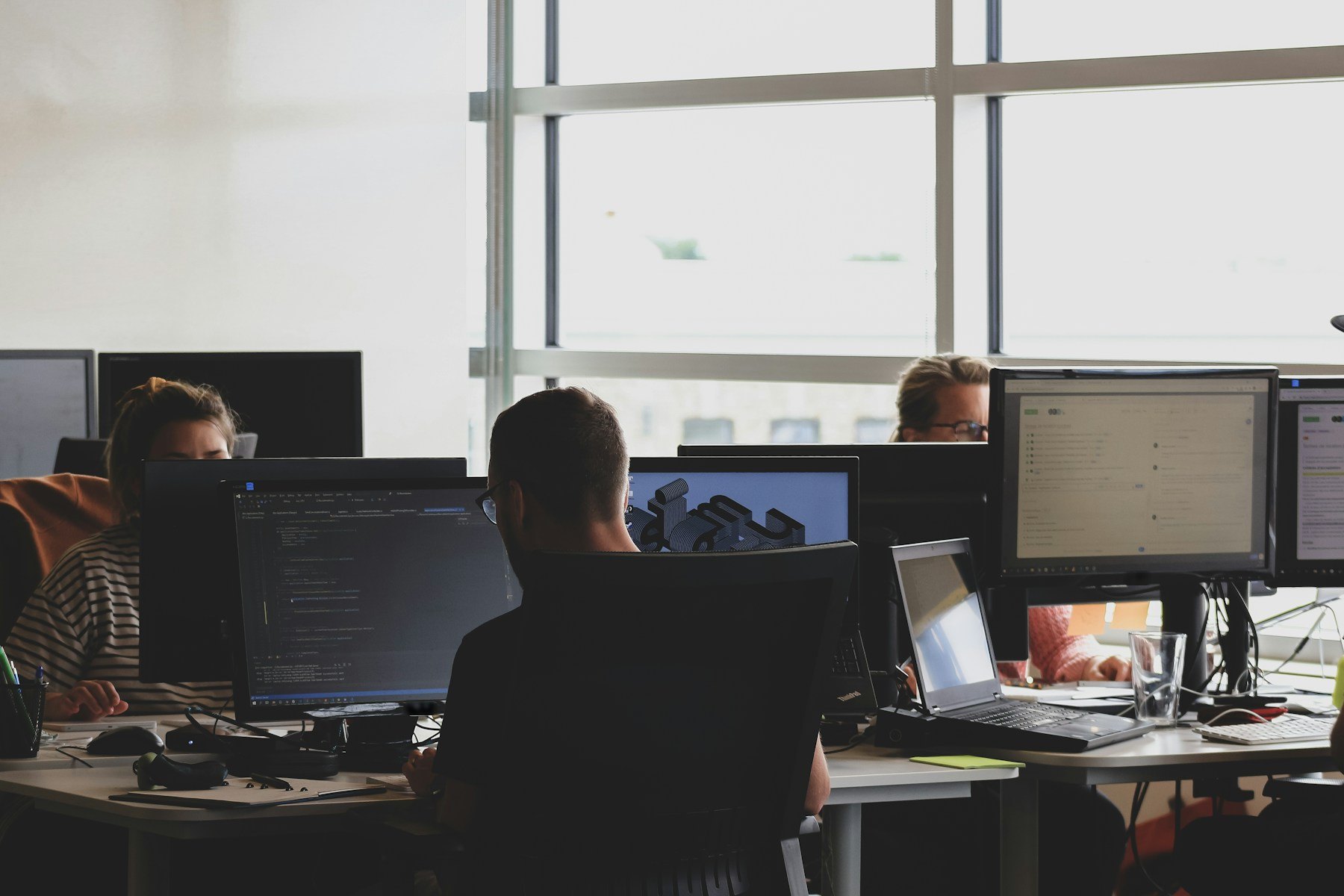 Three people work at a row of monitors in a bright office, one screen showing code and another a ticket board