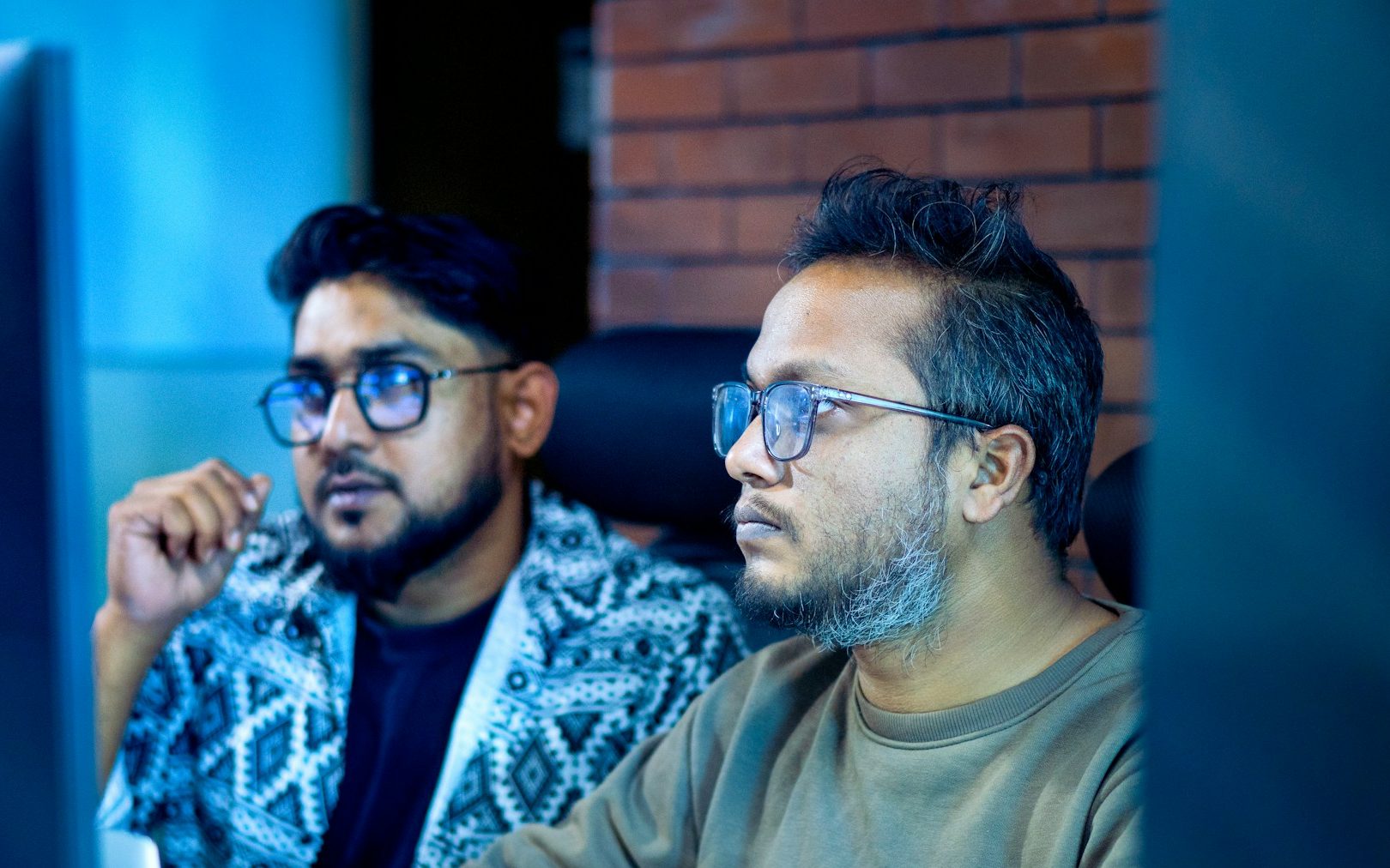 Two engineers in glasses sit side by side watching a monitor closely in a dim, blue-lit room, one gesturing as he talks