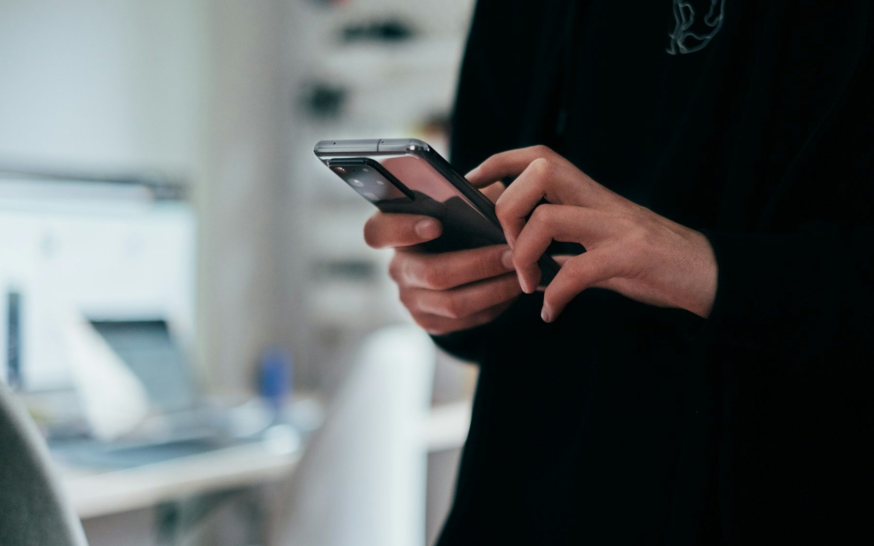 A person in a dark sweater taps a question into a smartphone held in both hands, a bright office blurred behind