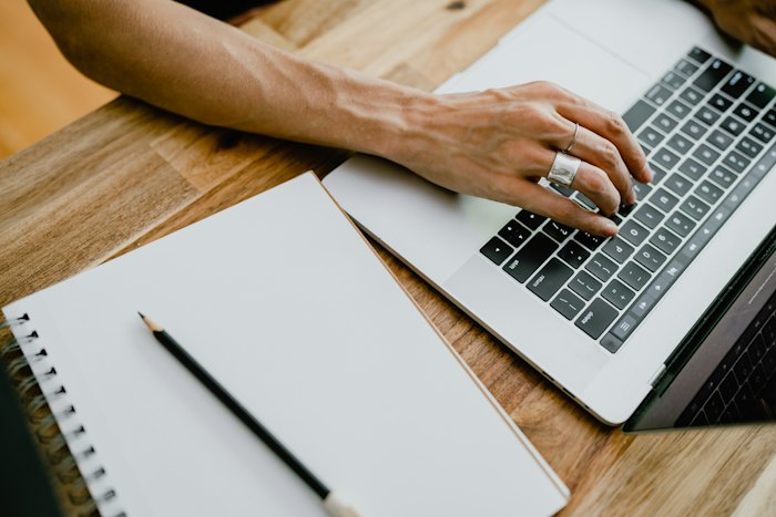 A hand typing on a laptop keyboard beside an open notebook and a pencil