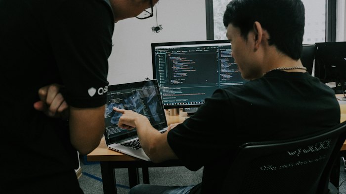 A developer pointing at a laptop screen of code while a colleague leans in