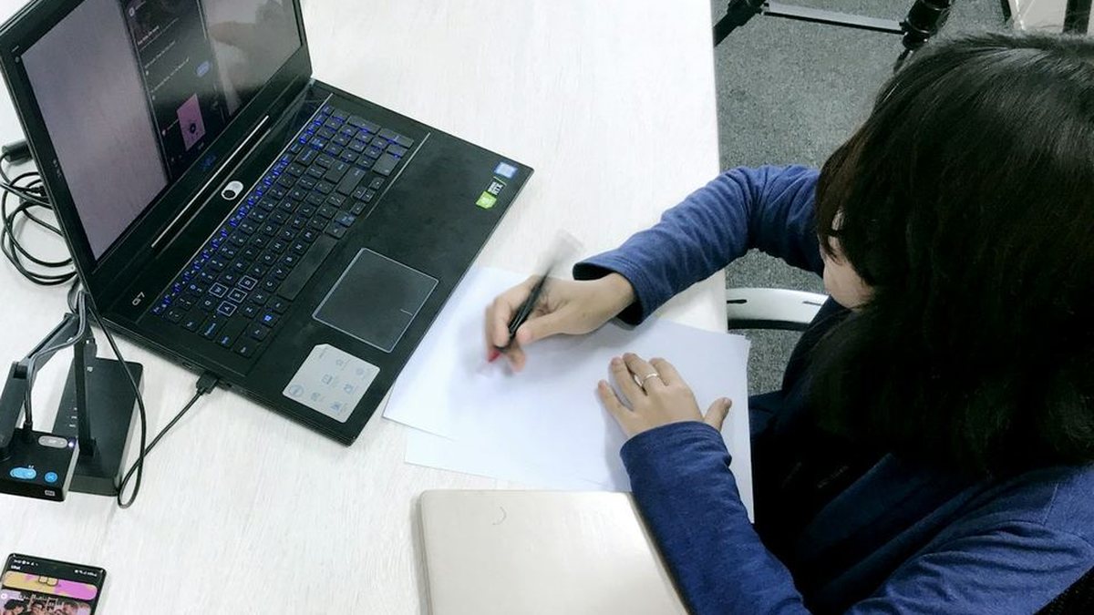 A researcher writing notes beside a laptop mirroring the phone screen a participant is using