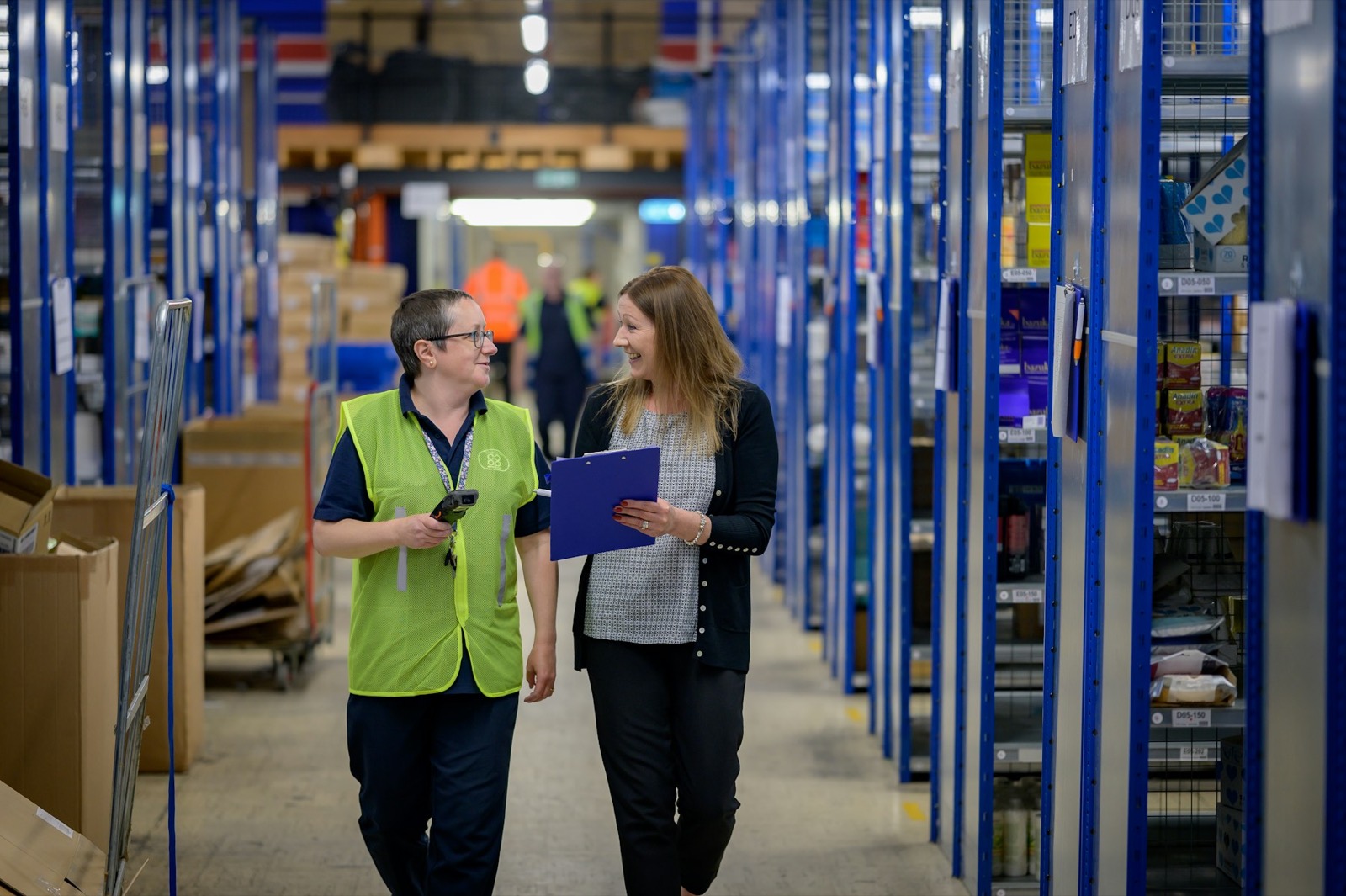 Two workers walking a warehouse aisle, one in a hi-vis vest holding a handheld scanner