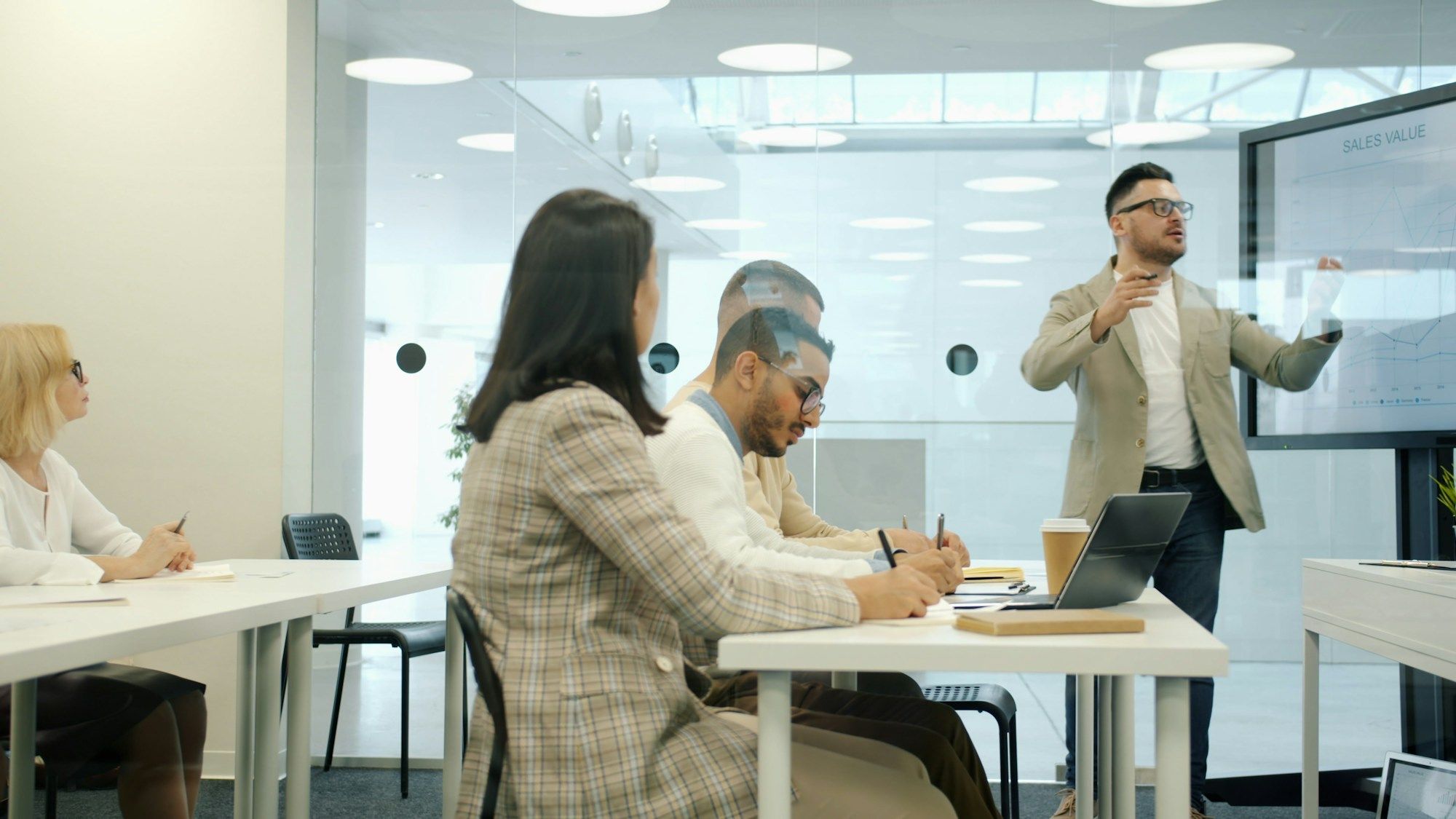 A man gestures beside a screen while colleagues take notes at white desks in a meeting room.