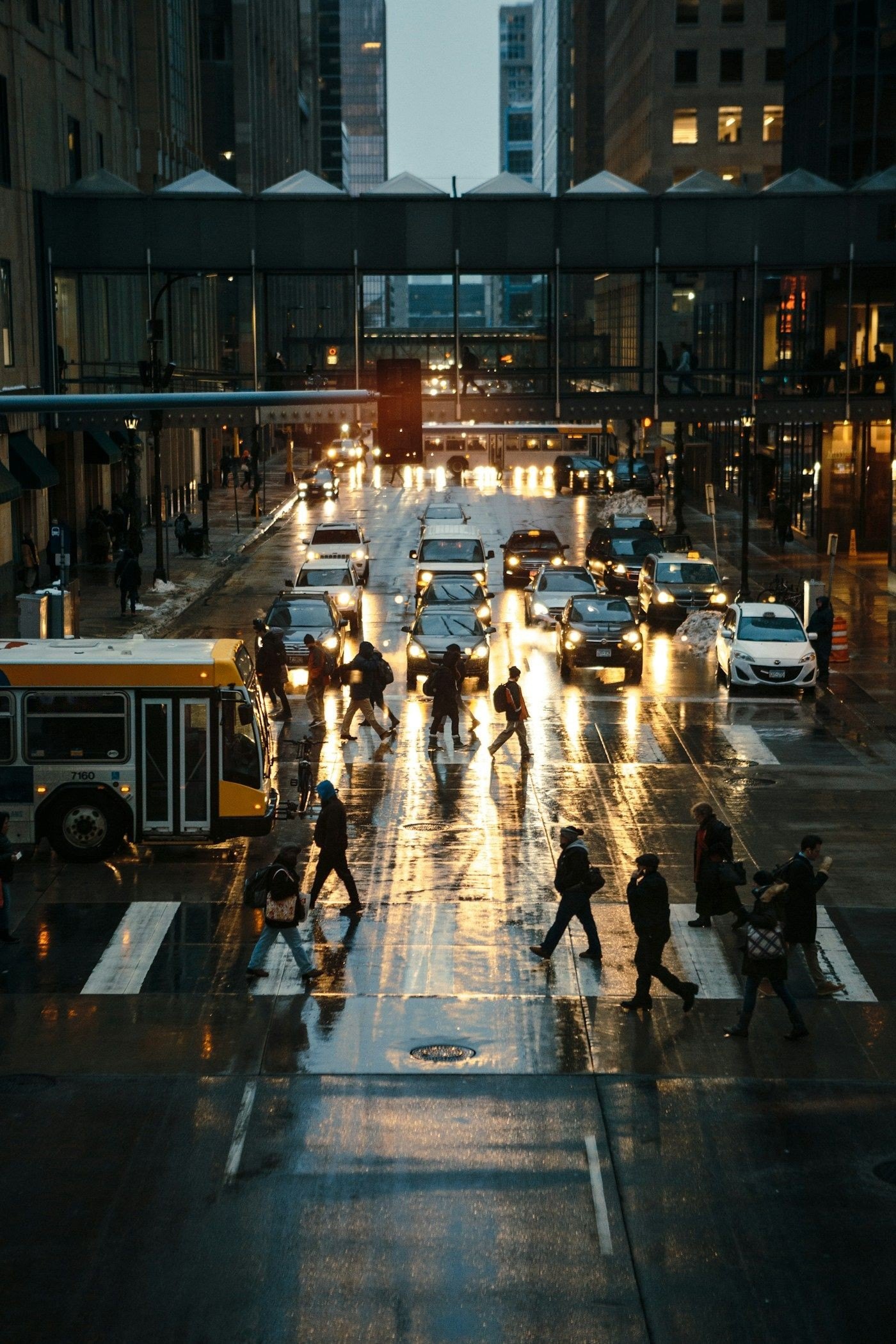 Pedestrians cross a wet city street at dusk, car headlights reflected on the road.