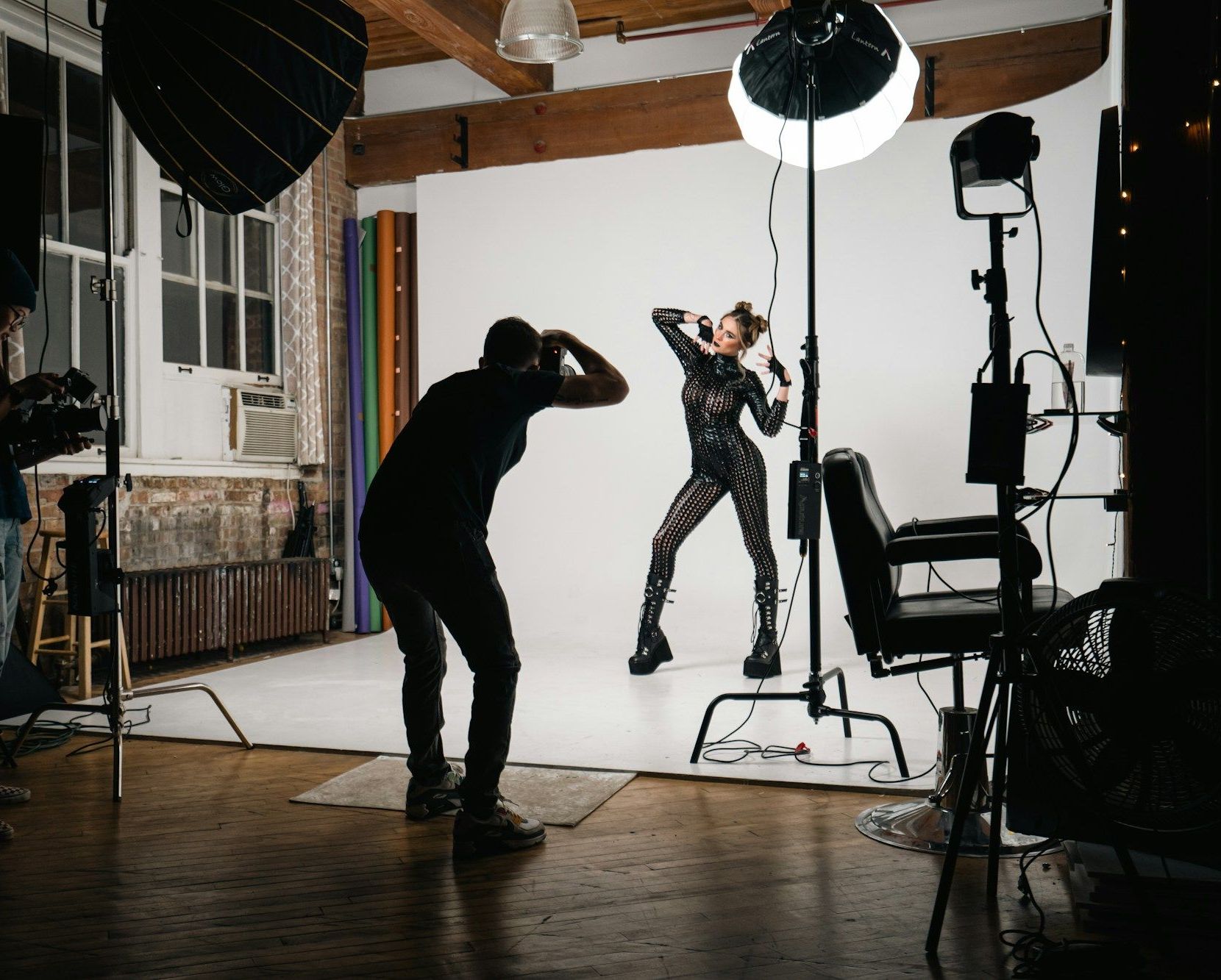 A photographer crouches to shoot a model in a black catsuit on a white studio set while a videographer films.